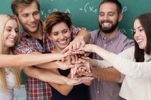 Teamwork and collaboration amongst students with a laughing group of diverse young men and women standing close together forming a stack with their hands-1